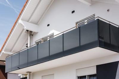 Anthrazitfarbenes Glasgeländer am Balkon eines weißen Hauses unter rotem Satteldach.